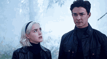 a man and a woman are standing next to each other in a forest looking at each other .