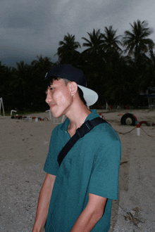 a young man wearing a green shirt and a black hat stands on a beach with palm trees in the background