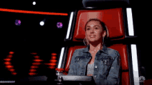 a woman in a denim jacket is sitting in a red chair with her hands folded and smiling .