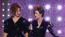 two drag queens standing next to each other in front of a purple curtain