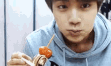 a young man in a blue hoodie is eating a skewer of food with a fork .
