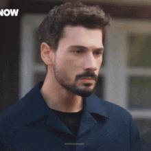 a man with a beard is standing in front of a sign that says " now "