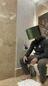 a man with a green hat on his head is sitting on a toilet
