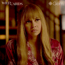 a woman with long blonde hair and bangs is wearing a red robe and a gem logo behind her