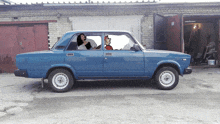 a blue car is parked in front of a garage with a woman sitting in the driver 's seat