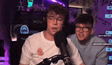 two young men are sitting in front of a microphone and one of them is wearing glasses .