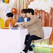 two men are hugging each other while sitting at a table with balloons in the background