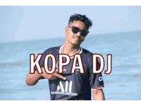 a man wearing sunglasses and a kopa dj shirt stands in front of the ocean