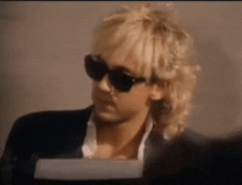 a man wearing sunglasses and a suit is sitting at a desk .