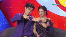 a man in a purple suit and a woman in a purple dress are sitting on a couch and making a heart shape