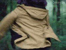 a woman is running through a forest wearing a tan jacket with a hood .