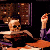 a skull sits on top of a stack of books next to a hand holding a cigarette