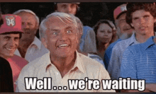 an older man is sitting in a crowd of people and smiling while saying `` well ... we 're waiting '' .
