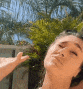 a woman is standing in front of a palm tree with her eyes closed and a hand reaching up towards her face .