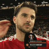 a man in a red shirt is talking into a microphone with the name mason mount on the screen behind him