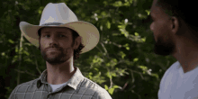 a man in a cowboy hat stands next to another man in a plaid shirt