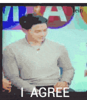 a man in a grey sweater says i agree in front of a tv screen