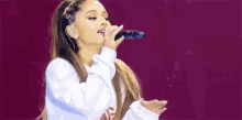 ariana grande is singing into a microphone on a stage while wearing a ponytail .