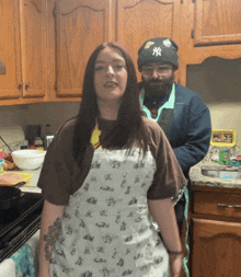 a man wearing a ny hat stands next to a woman wearing an apron in a kitchen