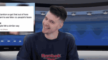 a man wearing a reebok sweatshirt is smiling in front of a screen that says attention so get that out of here
