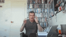a man is sitting in front of a laptop in front of a bookshelf full of dvds ..