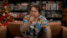 a man in a hawaiian shirt is sitting on a couch with a christmas tree in the background