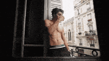 a shirtless man wearing lacoste underwear looks out of a window