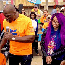a man in a yellow shirt is speaking into a microphone while a woman with purple hair stands next to him .