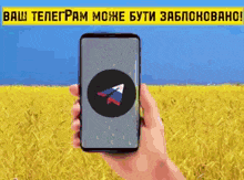 a person is holding a cell phone in front of a field with a banner that says " ваш телеграм може бути заблоковано "