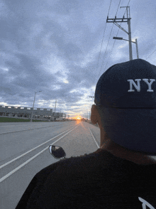 a man wearing a ny hat is driving down a road at sunset