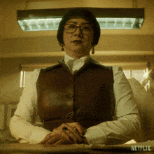 a woman wearing glasses and a brown vest is sitting at a desk with a netflix logo behind her