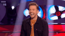 a man is laughing on a stage with a bbc logo in the background