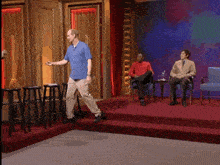 a man in a blue shirt stands on a stage while two men sit in chairs