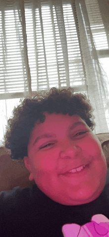 a young boy with curly hair smiles in front of a window with blinds