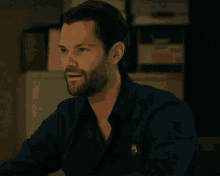 a man with a beard wears a blue shirt with a badge on his chest