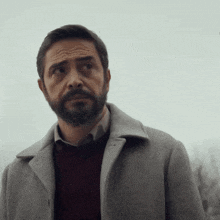 a man with a beard wears a grey coat and a red sweater