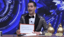 a man in a tuxedo is sitting at a table holding a sign that says ' certudo sottozero '