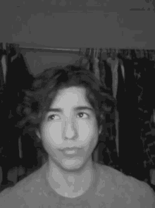 a black and white photo of a young man in front of clothes hanging on a rack