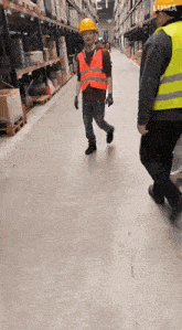 a man wearing a hard hat and safety vest is walking down a hallway in a warehouse .