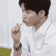 a young man wearing a white shirt and a necklace is eating something with his finger in his mouth .