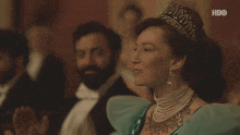 a woman wearing a tiara and pearls sits in front of a hbo sign