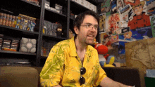 a man wearing glasses and a yellow shirt is sitting in front of a wall covered in pictures and books