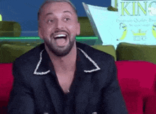 a man with a beard is sitting on a red couch with his mouth open and smiling .