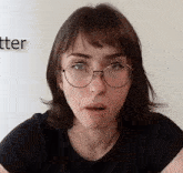 a woman wearing glasses and a black shirt with the word tter on the wall behind her