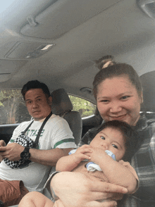 a man wearing a shirt that says ' the ' on it sits next to a woman holding a baby in a car