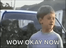 a young boy is standing in front of a car with the words wow okay now written on it .