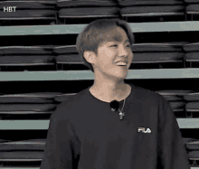 a young man is wearing a black fila shirt and smiling in front of a row of empty seats .