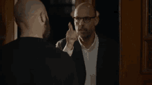 a bald man with glasses and a beard is pointing at his reflection in a mirror
