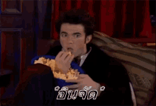 a man is sitting on a couch holding a bucket of popcorn with a foreign language behind him