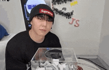 a young man wearing a supreme hat is sitting in front of a clear box filled with papers .